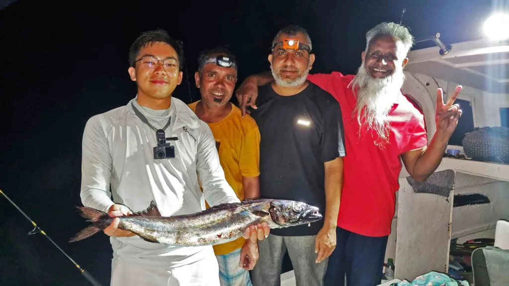 Anglers and local fishermen pausing for a photo while a tourist holds a freshly caught Kattelhi fish on a boat at night in Fuvahmulah.