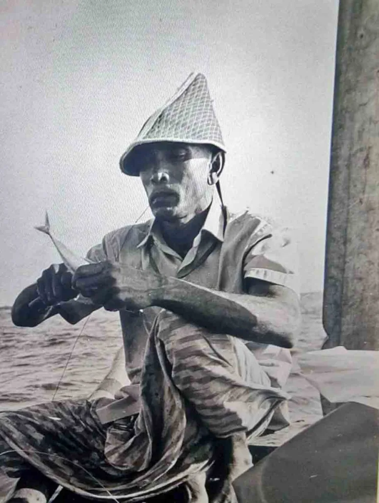 Black-and-white photograph of a Maldivian fisherman wearing a traditional woven hat, seated on a wooden vessel while carefully rigging a bait fish onto a line.
