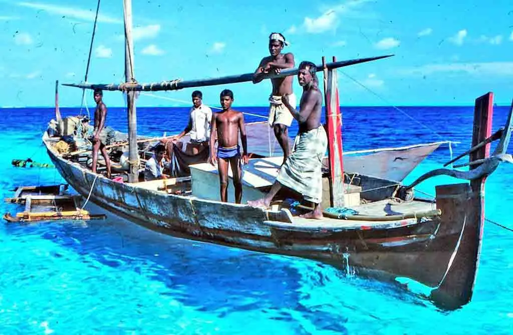 A group of Maldivian men and boys stand on a traditional wooden dhoani boat floating on vibrant turquoise water under a blue sky.